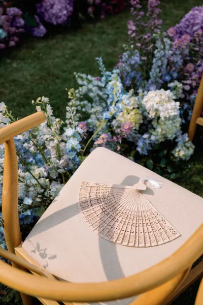 Wedding ceremony aisle florals with blue hydrangea aisle flowers in a lush garden, beside a wooden chair with a lace paper fan and tassel