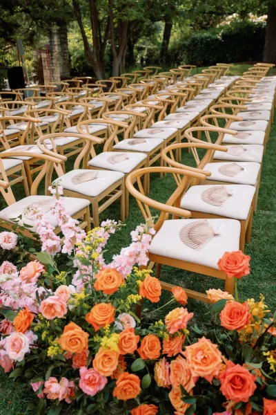 Ceremony seating with outdoor ceremony chairs, white cushions and handheld fans, beside colorful aisle flowers on a manicured lawn by hedges