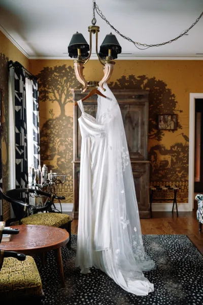 Wedding dress with veil hanging from a chandelier on a wooden hanger in a vintage bridal suite with patterned curtains and armoire