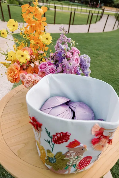 Wedding welcome basket with purple fans and colorful florals on a wooden table, set on a grassy lawn by a paved walkway and railing