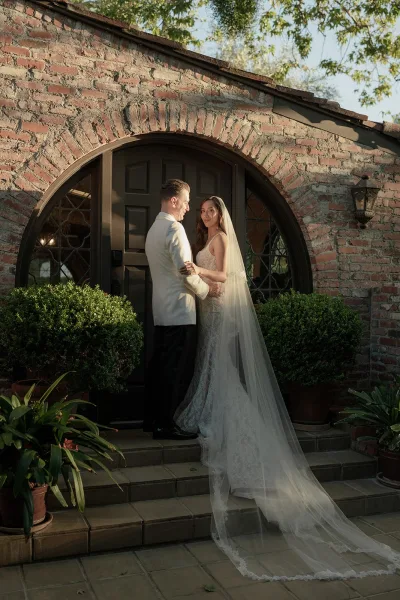 Couple portrait of the bride and groom on steps, her wedding veil trailing as they embrace by an arched brick doorway with leaded windows.