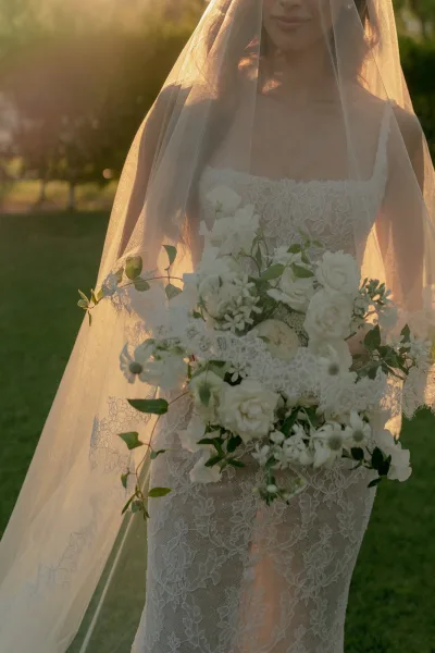 Bridal portrait of a bride under a veil with a white bouquet, standing amid garden trees on a grass lawn.