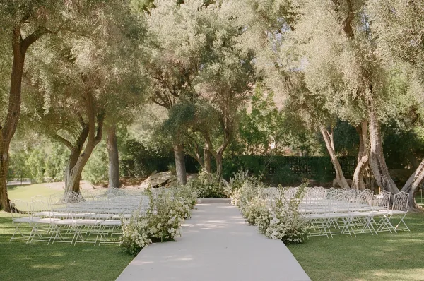Ceremony aisle design for an outdoor ceremony setup, bordered by rows of white metal chairs on a grass lawn under garden trees.