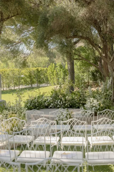Ceremony setup for an outdoor garden ceremony with white florals along the aisle, framed by garden trees and clipped hedges.