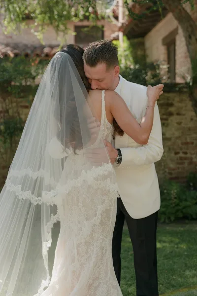 Wedding first look as bride and groom hug, her lace gown sweeping close, framed by garden greenery against a brick wall and clay tile roof.