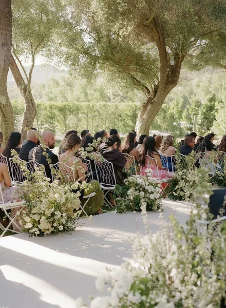 Ceremony aisle design featuring outdoor ceremony seating and aisle flowers on a grass lawn beneath trees, with hedges and distant hills.