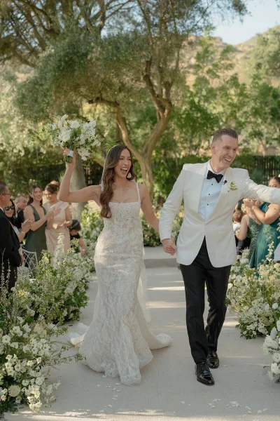 Ceremony recessional and just married exit as the couple passes cheering guests, the bride lifting a white and green bouquet in a lush garden.