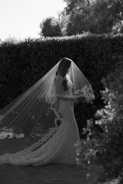 Bridal portrait of a bride with veil in a lace gown, standing on a stone path framed by garden hedges and trees.
