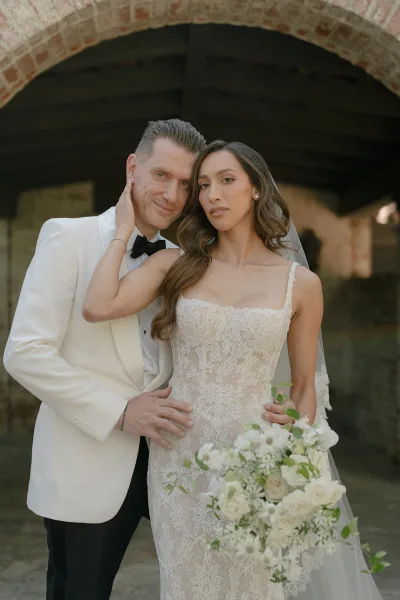 Couple portrait of a bride in a lace gown and a groom in a white tuxedo beneath a brick arch, her white rose bouquet bright against the rustic wall.