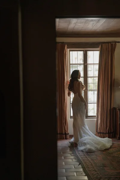 Bridal portrait of a bride by window, poised at a wrought iron window in a rustic room with plaster walls and terracotta tile floor.