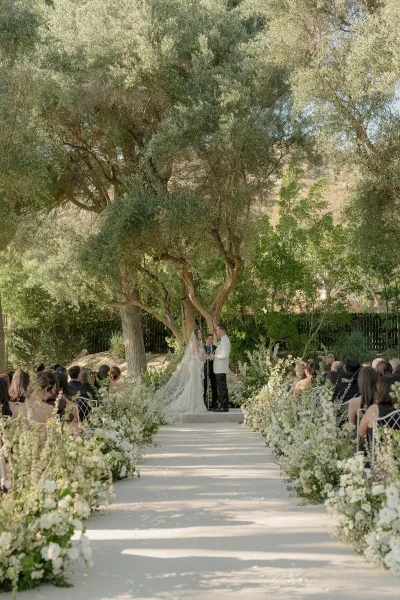 Ceremony moment at an outdoor wedding ceremony as the couple stands beneath trees, with aisle flowers lining a garden path.