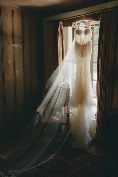 Wedding dress hanging by a window, a lace wedding dress with a veil draped over the wood chair beside it on a patterned rug