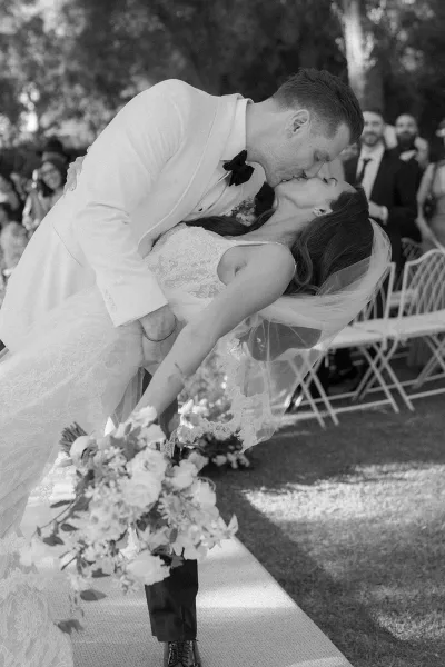 Wedding kiss portrait capturing a dip kiss moment as the groom dips the bride, her bridal bouquet lifting, with guests beneath garden trees.
