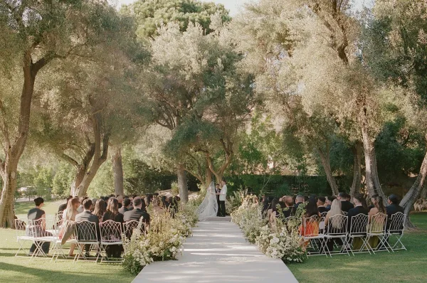 Ceremony moment during an outdoor wedding ceremony as the couple stands on a white aisle runner beneath trees on a grass lawn.