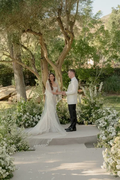 Ceremony moment during an outdoor wedding ceremony as the couple exchanges vows, her veil catching light beneath garden trees with a hillside beyond.