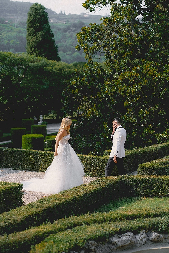 First look moment as bride in strapless gown with long train and tiara approaches groom in tuxedo on a gravel path in a formal garden