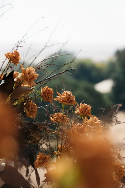 Wedding floral arrangement with orange rose wedding flowers, dried branches and grasses, set outdoors with trees, sky, and distant water