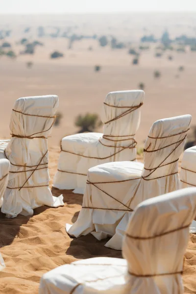 Ceremony seating with white chair covers and rope ties arranged on sand, set against desert dunes, sparse shrubs, and open sky