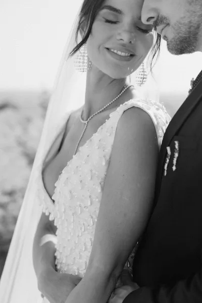 Couple portrait of bride and groom close up, embracing cheek to cheek as he kisses her forehead, veil and beaded gown against open sky
