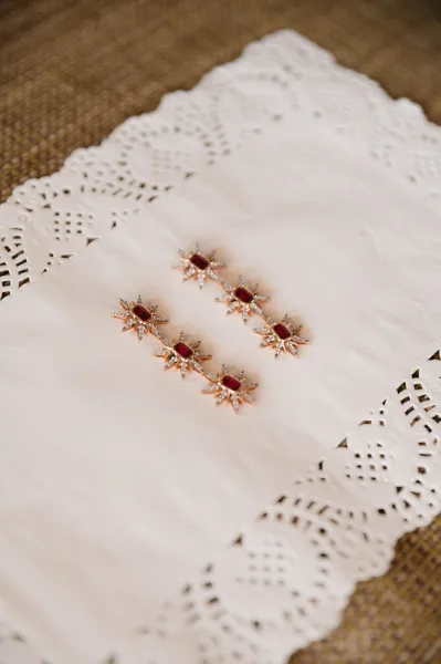 Bridal jewelry wedding earrings with red gemstone and crystal accents, arranged beside a lace handkerchief on woven fabric surface