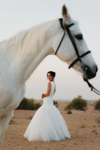 Bridal portrait of a bride with horse, wearing a beaded low-back tulle gown and necklace, looking over her shoulder in a desert landscape