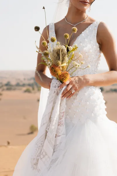 Bridal bouquet of dried wildflowers and protea with lace wrap, held against a beaded gown and veil in an open desert sky backdrop