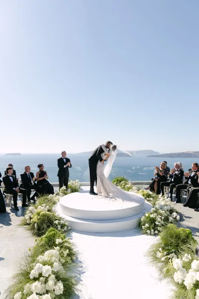 Wedding first kiss at an ocean view ceremony, couple embracing on a round ceremony platform with cliffs and blue sea beyond a glass railing.