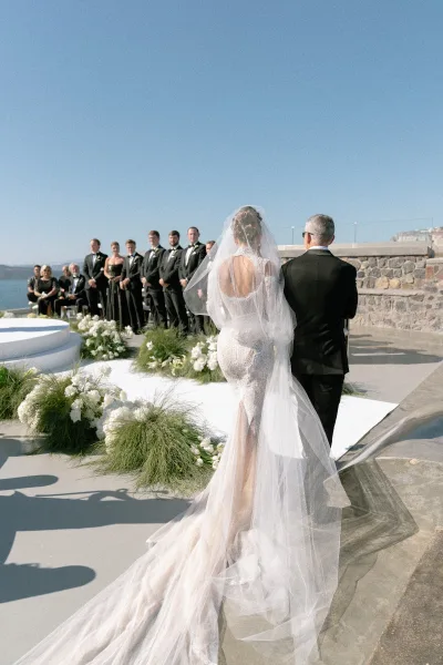 Wedding processional with a bride walking down the aisle, her long train trailing toward the altar on an oceanfront terrace under blue sky.