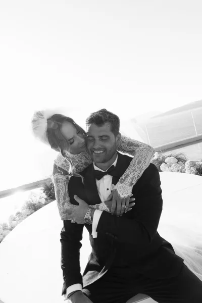 Couple portrait of a bride hugging groom, her lace wedding dress brushing his shoulder, on an ocean-view terrace with glass railing and hills.