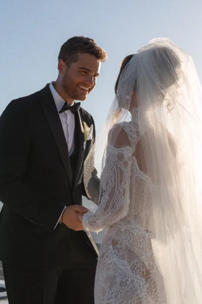 Ceremony moment during wedding vows as the bride and groom hold hands, her lace wedding dress catching light beneath a blue sky.