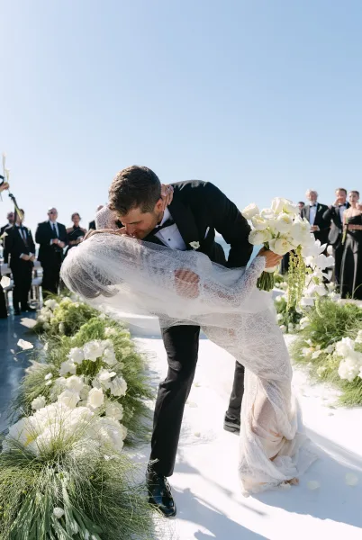 Ceremony kiss moment becomes a recessional kiss as the groom dips the bride, her veil sweeping the white aisle runner under a blue sky.