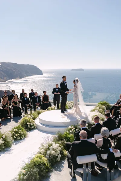 Ceremony moment as the couple holds hands during an ocean view wedding ceremony, framed by white florals, with sunlit cliffs and sparkling water.