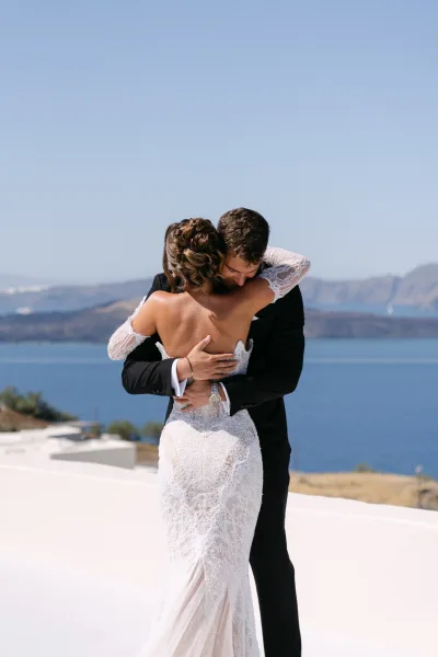 Couple portrait as the bride and groom hug on a white terrace, her off-the-shoulder sleeves delicate against blue sky, ocean, and coastal cliffs.