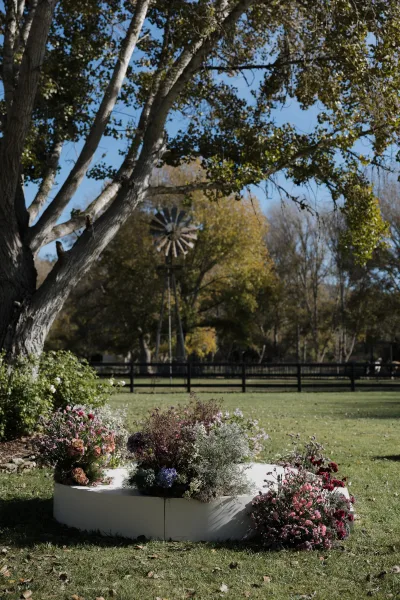 Wedding ceremony altar on a white platform with ground wildflower blooms, set on an outdoor lawn with trees and a windmill backdrop