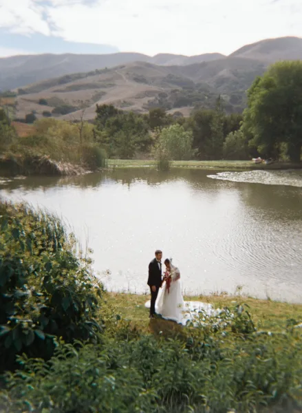 Couple portrait of bride in veil with bouquet and groom in tuxedo on a lakeside shore, mountains and cloudy sky behind