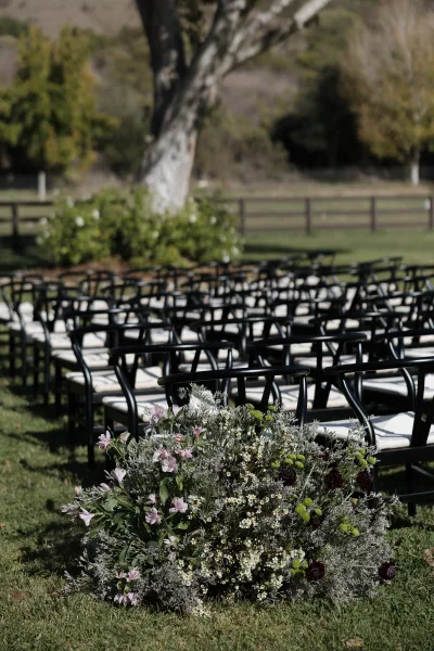 Ceremony seating with black chairs on a grass lawn, rows facing an aisle with wildflower accents, trees and rolling hills beyond