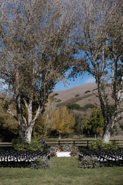 Ceremony setup with black chairs and a white aisle runner leading to a floral platform on a green lawn beneath large trees and hills