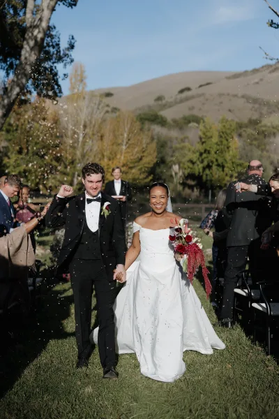 Wedding recessional as newlyweds walk hand in hand through ceremony exit confetti, bride in veil holding bouquet on sunny lawn aisle