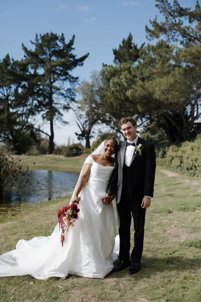 Couple portrait of bride and groom holding hands by a pond, bride leaning on groom with veil and red cascading bouquet under blue sky