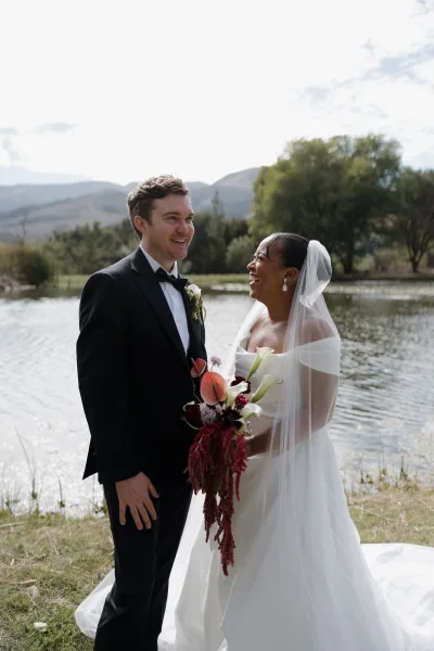 Couple portrait of bride and groom laughing, bride holding calla lily bouquet with veil, by a lakeside with mountains and trees