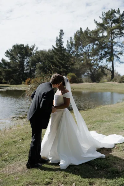 Wedding kiss portrait of bride and groom kiss by a lake, her off-the-shoulder gown and long veil trailing on the grass near reeds