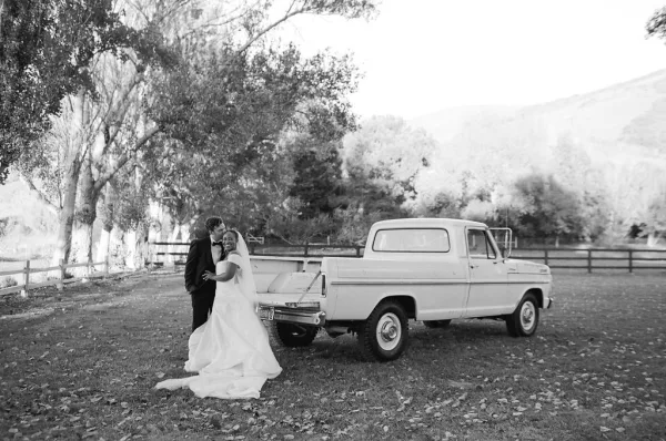 Couple portrait in a black and white wedding photo, bride in long veil and train with groom in bow tie beside a vintage pickup truck.