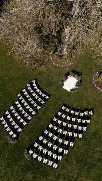 Outdoor ceremony setup with a white ceremony platform and floral arrangements, surrounded by curved chair layout on a garden lawn under trees