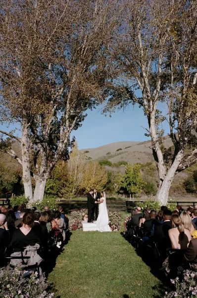 Wedding ceremony outdoors with bride and groom at a floral arch, aisle flowers leading to the altar on a lawn with mountains beyond