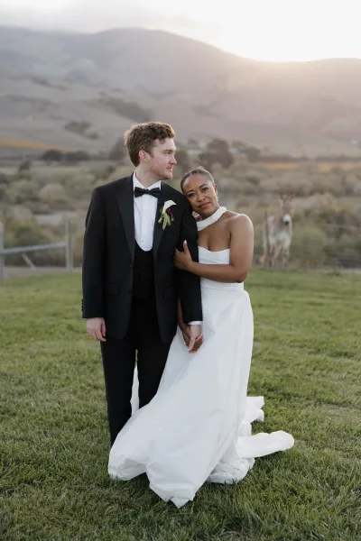 Couple portrait of bride in a simple wedding dress and groom in tuxedo holding hands in a grassy mountain field at sunset light