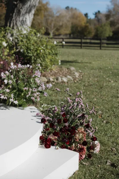 Ceremony aisle decor with wedding aisle flowers in burgundy, pink, and purple lining a white platform on a grassy lawn by trees and fence