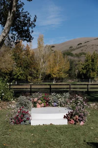 Ceremony aisle decor with a white runner and floral lined aisle of roses, hydrangeas, and greenery on a grassy lawn by a wooden fence