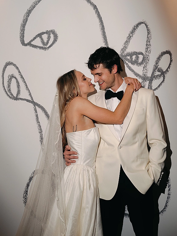 Couple portrait of bride and groom embrace, forehead touching, her strapless dress and veil against a white wall with abstract line art mural