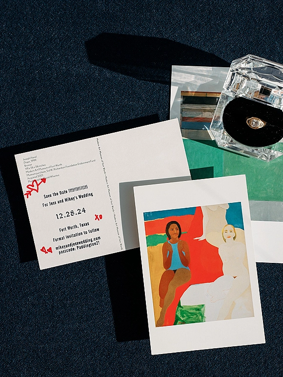 Save the date card in a modern save the date flatlay with red doodles, engagement ring in clear acrylic box, on a dark textured surface