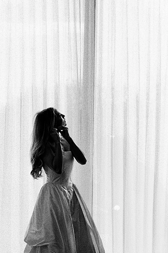 Bridal portrait of a bride getting ready, adjusting earrings in a strapless ball gown by a window with sheer curtains in soft light